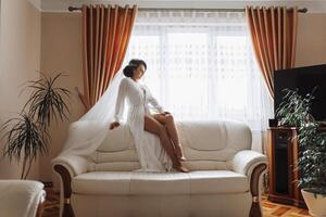 un increíblemente hermosa novia en el Mañana en un blanco túnica y un largo velo se sienta en el cama en su dormitorio. el novia poses en el Mañana antes de el Boda ceremonia. foto