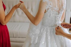 The bride's friend helps her get dressed in a white wedding dress in the morning. wedding morning, blurred focus. A friend of the bride helps to tie her wedding dress. photo