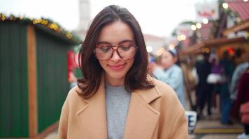 une femme portant des lunettes et une manteau permanent dans une bondé endroit avec flou Noël lumière à le Contexte est à la recherche à sa téléphone video