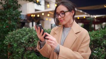 een vrouw in bril en jas is op zoek Bij haar telefoon in buitenshuis boodschappen doen wijk video