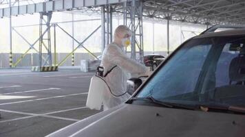 un hombre en un protector traje y máscara con un agua pulverización herramienta es desinfectante un coche video