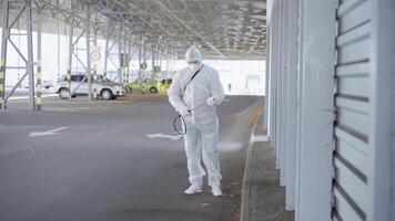 un hombre en un protector traje y máscara con un agua pulverización herramienta es pulverización el desinfectante en el estacionamiento lote video