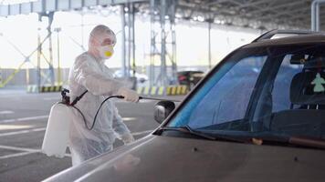 une homme dans une protecteur costume et masque avec une l'eau pulvérisation outil est désinfectant une voiture dans le parking lot video