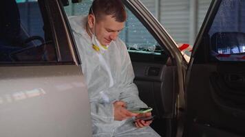 un hombre vistiendo su protector traje es sentado en su coche mientras mirando a su teléfono en un vacío estacionamiento lote video