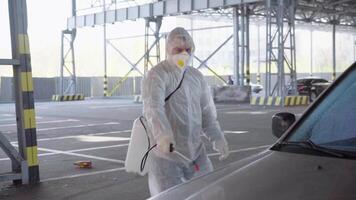 un hombre en un protector traje y máscara con un agua pulverización herramienta es desinfectante un coche en el estacionamiento lote video