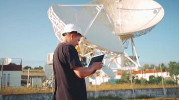 hombre en un difícil sombrero es trabajando en un tableta en frente satélite platos al aire libre video