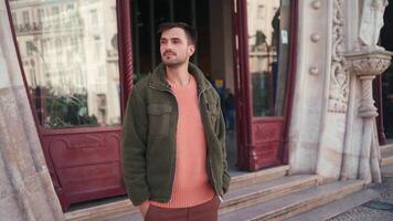 a man in a green jacket is standing in front of a building, looking at his phone and waving video