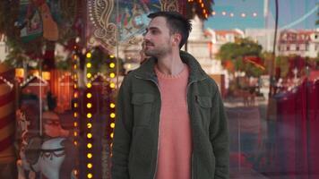 a man in a green jacket is standing in front of a carousel is facing left then smiling to the camera video