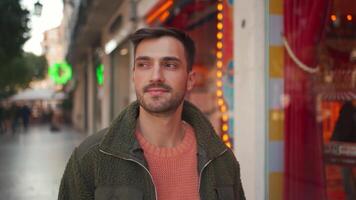 portrait de une Jeune homme portant vert manteau permanent dans de face de une boutique dans une achats district souriant à le caméra video