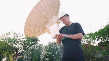 een Mens in een moeilijk hoed werken Aan zijn laptop terwijl staand in voorkant van een satelliet schotel video