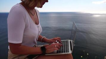 digital nômade, o negócio mulher trabalhando em computador portátil de a mar. bonita senhora digitando em computador de a mar às pôr do sol, faz uma o negócio transação conectados a partir de uma distância. freelance, controlo remoto trabalhos em período de férias video