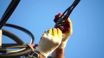 Skilled electrician in helmet fixes wires standing on ladder near high pole against blue sky on summer day backside view. Electrical service and mounting on the pole. Slow motion video