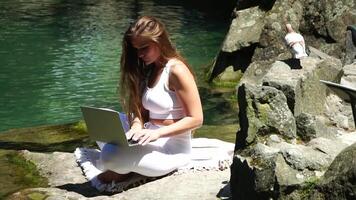 bene guardare mezzo anziano donna digitando su il computer portatile tastiera all'aperto vicino lago nel il parco. vicino su donna mani scrittura su computer. libero professionista, digitale nomade, viaggio e vacanze concetto. video