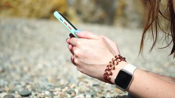 Woman with smartphone. Close-up of woman's hands holding vertical mobile phone and swiping up finger application page against background of sea and beach video