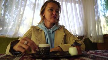 Woman pouring Turkish coffee from cezve into cup. Closeup slow motion shot of female hand with cup on square plate, on table in cafe outdoor. Traditional hot unfiltered coffee served in restaurant video