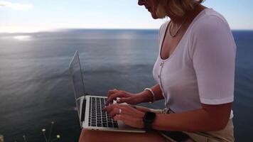 digital nômade, o negócio mulher trabalhando em computador portátil de a mar. bonita senhora digitando em computador de a mar às pôr do sol, faz uma o negócio transação conectados a partir de uma distância. freelance, controlo remoto trabalhos em período de férias video