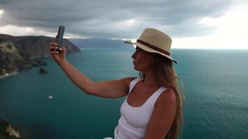 Woman travel sea. Happy tourist in hat enjoy taking picture outdoors for memories. Woman traveler posing on the beach at sea surrounded by volcanic mountains, sharing travel adventure journey video