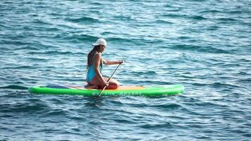 hav kvinna supera. silhuett av Lycklig positiv ung kvinna i blå bikini, surfing på grön supera styrelse genom lugna vatten yta. idyllisk solnedgång. aktiva livsstil på hav eller flod. långsam rörelse video