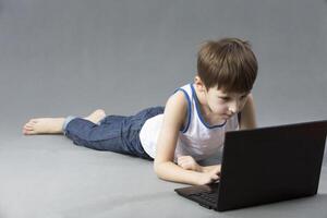 niño con un computadora. el chico es acostado en el piso con un ordenador portátil foto