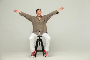 A middle-aged woman in casual clothes and red sneakers poses with a smile in the studio against a gray background with her arms outstretched. photo