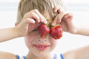 un niño con fresas el chico sostiene dos rojo bayas cerca el ojos foto