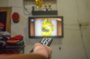 The hand is holding the TV remote in front of a television, preparing to change the TV channel photo