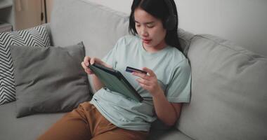 Footage high angle view shot, Happy young woman wear wireless headphones sitting on sofa using digital tablet and credit card for online shopping cashless in living room at home video