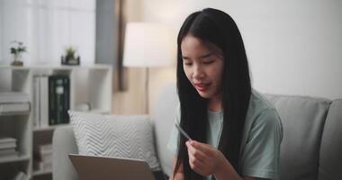 imágenes estable disparo, joven asiático mujer sentado en sofá participación crédito tarjeta haciendo en línea pago para compra en web Tienda utilizando ordenador portátil. comercio electrónico sitio web pagos concepto. video