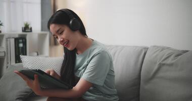 imágenes muñequita disparo, contento joven mujer vestir inalámbrico auriculares sentado en sofá y utilizando digital tableta para en línea compras sin efectivo en vivo habitación a hogar video