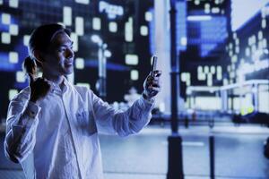 Man outside in city at night, using mobile phone to take selfie. Citizen using smartphone to take pictures of himself showing ok hand gesture sign while on empty streets illuminated by lamps photo