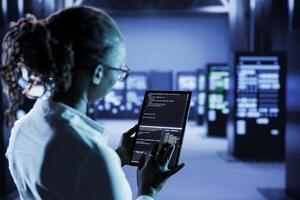 Computer scientist builds firewall protection systems in server room housing advanced infrastructure. High tech facility manager runs code on tablet to upgrade data center rigs photo