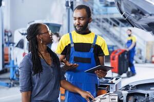 Mechanic at auto repair shop conducts annual vehicle checkup, informing customer about needed motor oil replacement. BIPOC garage worker talks with customer after finishing car inspection photo