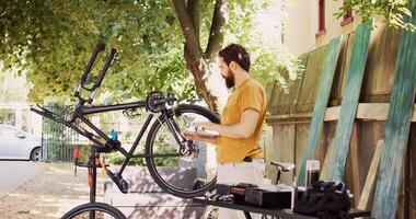 Male cyclist holding personal computer and inspects bike pedal and crank arm for damages in home yard. Active caucasian man enjoys browsing internet, fixing and maintaining modern bicycle. photo