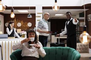 Friendly african american bellboy assists senior man in hotel reception with luggage to reserved room. Elderly female visitor sipping tea while waiting for check-in in comfy lounge area with sofas. photo