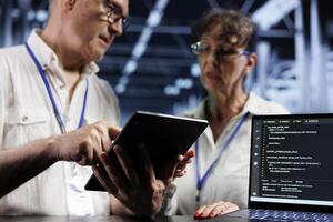 Octogenarian cloud computing company executives updating blade servers, writing intricate binary code scripts on tablet and laptop, using programming to fortify cybersecurity in data center photo