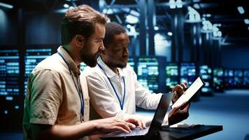 Coworkers running data center hardware diagnostic tests to determine and service software issues. Servicemen using laptop and tablet to fix critical systems, checking for blips photo