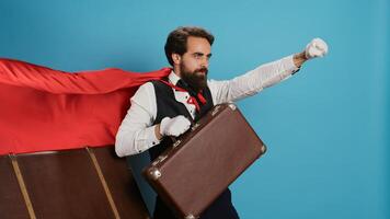 Hotel concierge acting like superhero with red cape, offering to help people with suitcases on camera. Young adult doorman presenting comic character with mantle, feeling determined and confident. photo