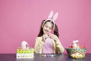 contento alegre niño con conejito orejas pintura huevos en preparación para Pascua de Resurrección domingo adornos, colorante decoraciones hecho a mano como un tradición. alegre niña decorando con acuarela teñir. foto