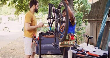 Committed interracial couple outside servicing bicycle components. Healthy active black woman and caucasian man inspecting and maintaining bike tires and chains for leisure cycling. Pull-back shot. photo