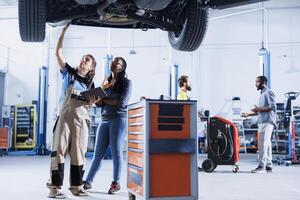 Adept mechanic in garage using laptop to follow checklist while doing maintenance on car, talking with client. Expert in auto repair shop does checkup on vehicle tires next to customer photo