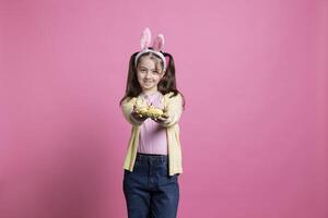 Sweet young girl confidently smiling for the camera while carrying a basket full of Easter eggs and a chick. Joyful toddler with rabbit ears, pigtails and spring ornaments stands over pink backdrop. photo