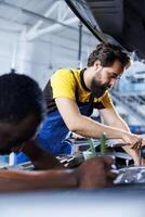 Repairman and coworker collaborating on servicing broken vehicle, checking for faulty motor. Licensed mechanics in auto repair shop working together on fixing car, discussing best options photo