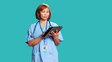 Licensed nurse wearing protective scrubs imputing medical data on tablet. Healthcare specialist happy at work, typing on digital screen, isolated over blue studio background photo