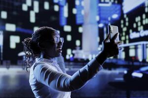 African american woman using mobile phone to take selfie while walking around city at night. Citizen using smartphone to take pictures of herself while strolling outside on empty streets photo