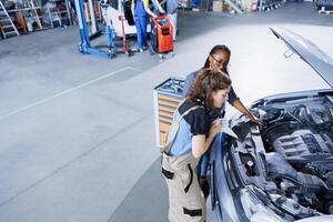 Skilled technician in garage finishing fixing car for BIPOC woman, looking underneath vehicle hood to remove remaining oil leaks. Worker does routine ignition system cleaning on client automobile photo