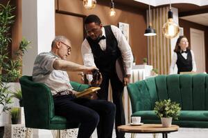 Retired old man pays with credit card at POS in hotel lobby, assuring quick and secure reservation transaction. Elderly guy with book makes cashless payment with help from african american waiter. photo