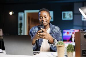 African american lady with positive emotion, using mobile device to surf the internet and chat with friends. Smiling black woman sitting in front of laptop, browsing social media on her smartphone. photo