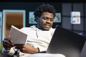 Joyful freelancer working from apartment, writing down business ideas while watching online video. Smiling man using pen and paper to note strategy plan, learning from internet clip on laptop photo