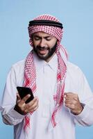 Excited man wearing traditional muslim outfit checking good news message while using smartphone. Happy cheerful muslim person showing winner gesture while scrolling internet page on mobile phone photo