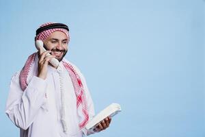 Smiling muslim man dressed in traditional outfit speaking on landline phone and having fun conversation. Arab person holding retro telephone and answering call with cheerful expression photo